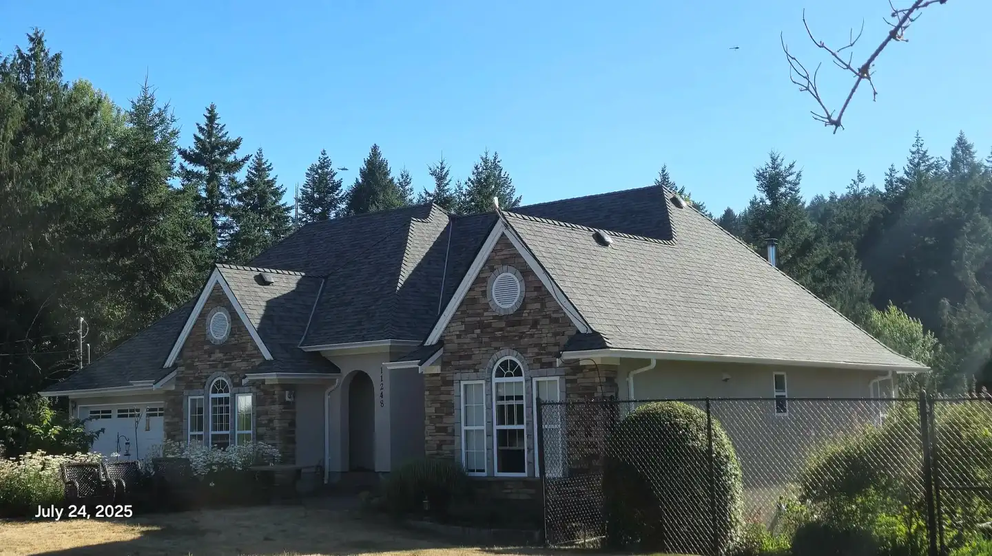 A steep-roofed house with a stone facade, surrounded by trees — a recent Leading Edge Roofing project on Vancouver Island.