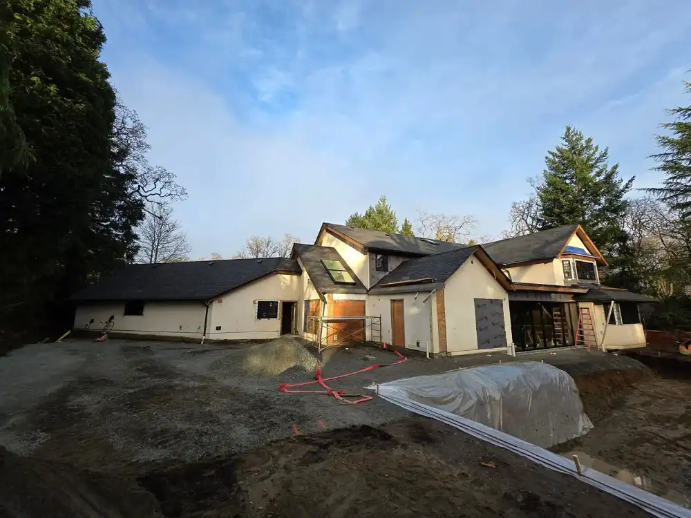 House under construction with scaffolding and exposed dirt, ringed by trees under a cloudy blue sky.